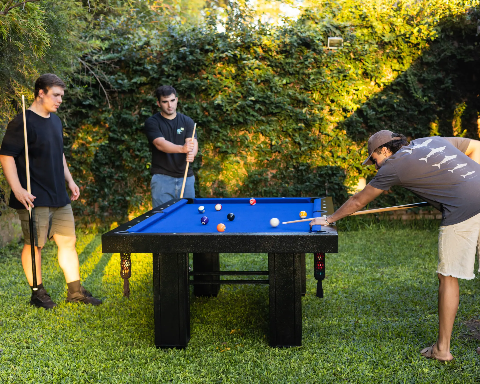 Tres amigos jugando al pool en una mesa BlackSteel® de paño azul, en un jardín