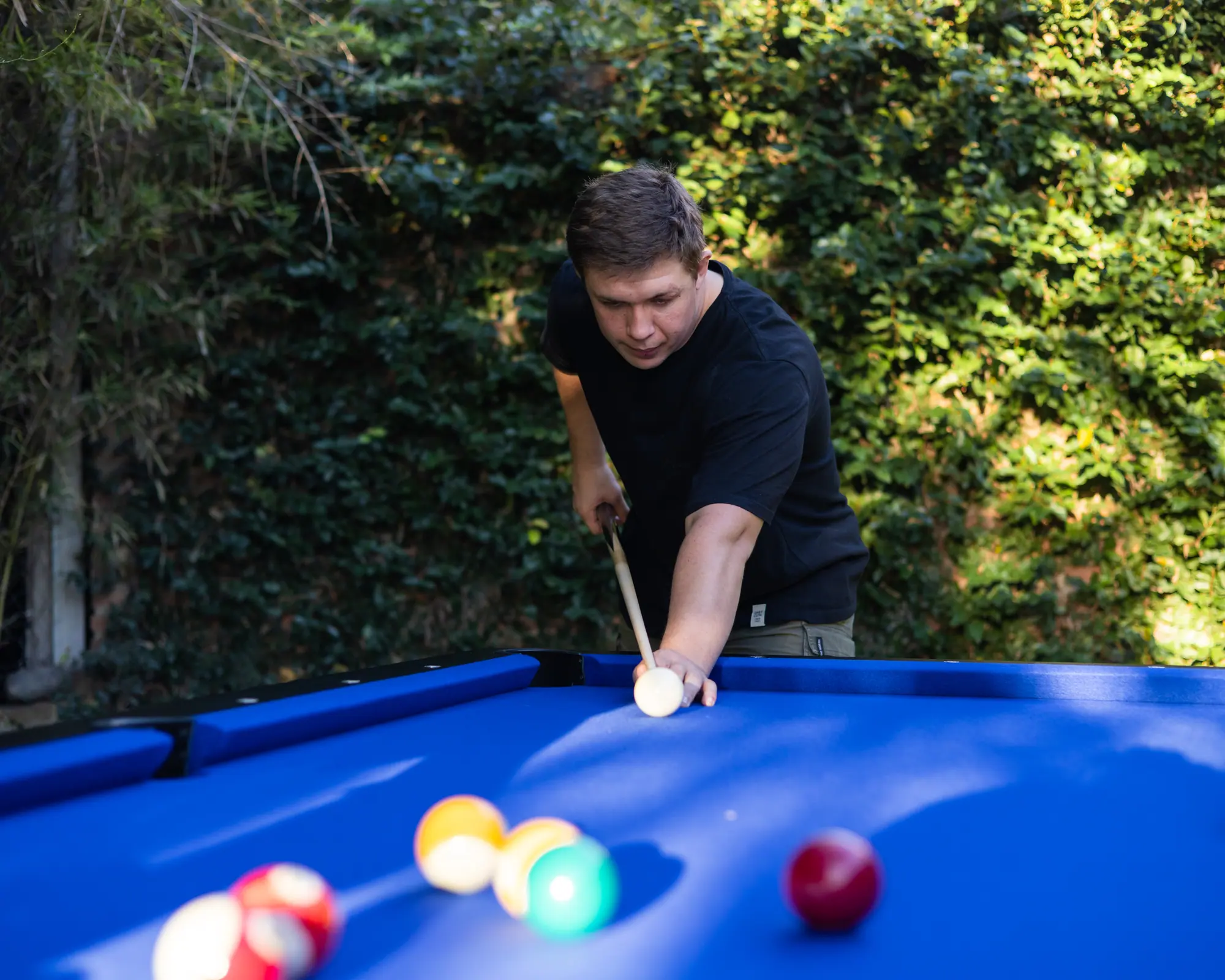 Jugador apuntando con el taco sobre una mesa BlackSteel® de paño azul, en un jardín